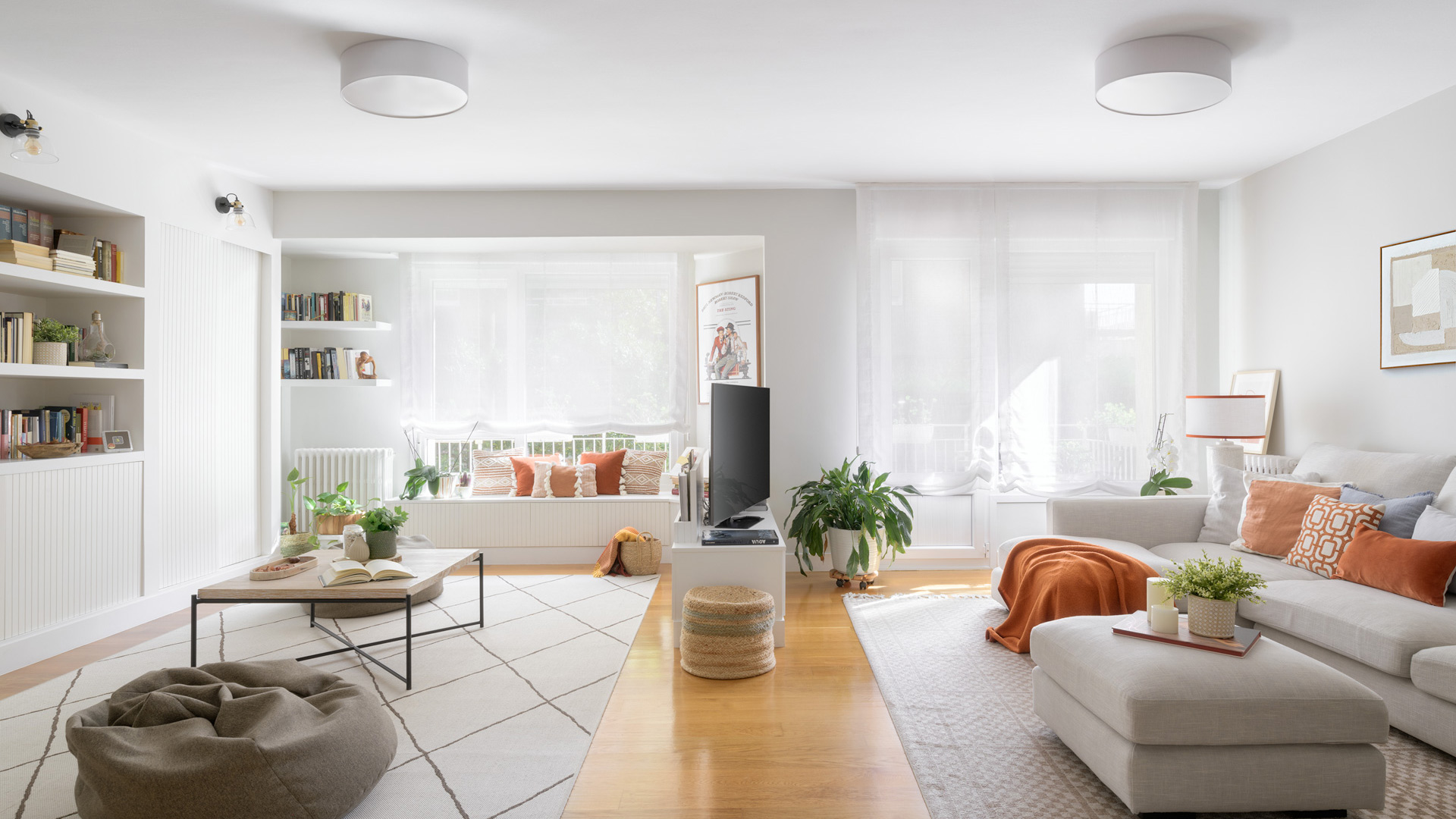 Sala de estar moderna con sofá beige, cojines naranjas, otomana central y plantas decorativas.